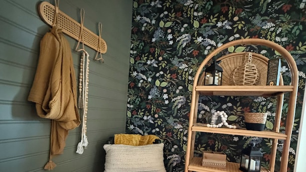 Una habitación con pared verde oscuro, papel tapiz floral, una silla negra, un estante de mimbre y un perchero con un paño marrón y un collar de cuentas.