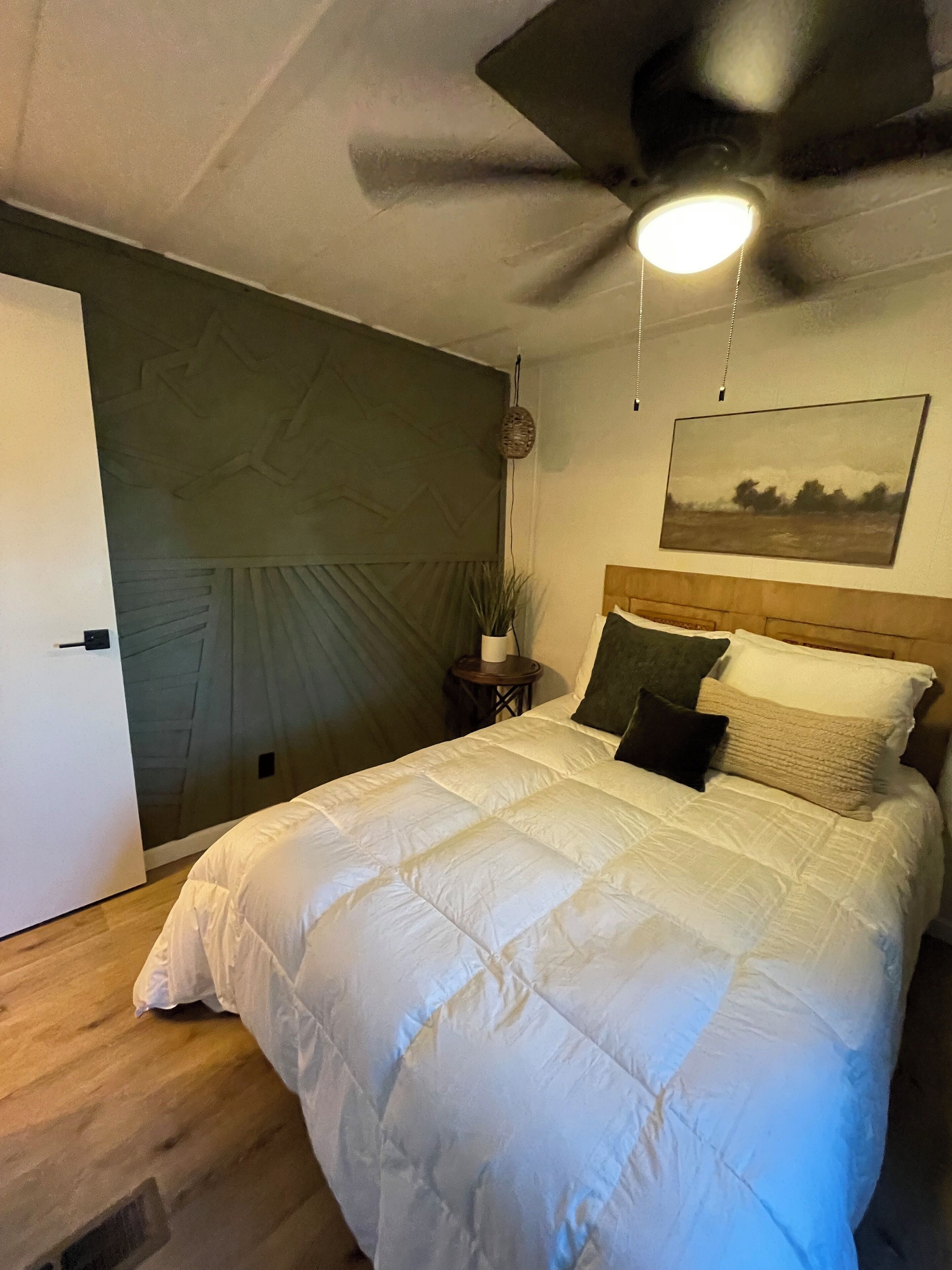 Ein Schlafzimmer mit einem Bett, einem Deckenventilator, einer Wandleuchte, einem kleinen Tisch mit einer Pflanze und einem Bild an der Wand.