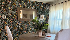 Un comedor con una mesa redonda de madera, dos sillas tapizadas, una planta en maceta y una pared con estampado floral.