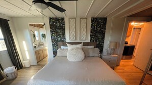 Un dormitorio con una cama grande, un ventilador de techo, una mesita de noche, un aparador y un baño al fondo.