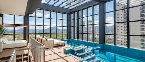 A rooftop infinity pool with a glass ceiling, wooden deck, and a seating area with a view of the cityscape.