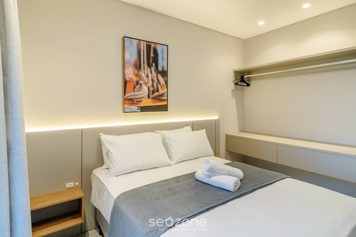 A neatly made bed with white linens, a gray blanket, and a rolled towel.