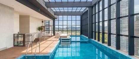 An indoor swimming pool with a wooden deck, a glass ceiling, and a view of the cityscape.