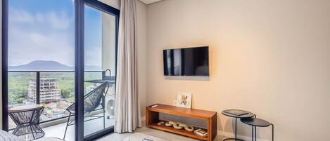 Uma sala de estar moderna com uma grande porta de vidro deslizante que dá para uma varanda, uma TV de tela plana na parede, uma mesa de console de madeira e um tapete estampado.