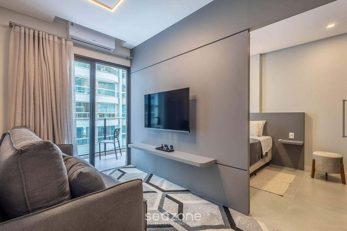 A modern hotel room with a sofa, a TV mounted on a wall, a bed, a small table, and a window with curtains.