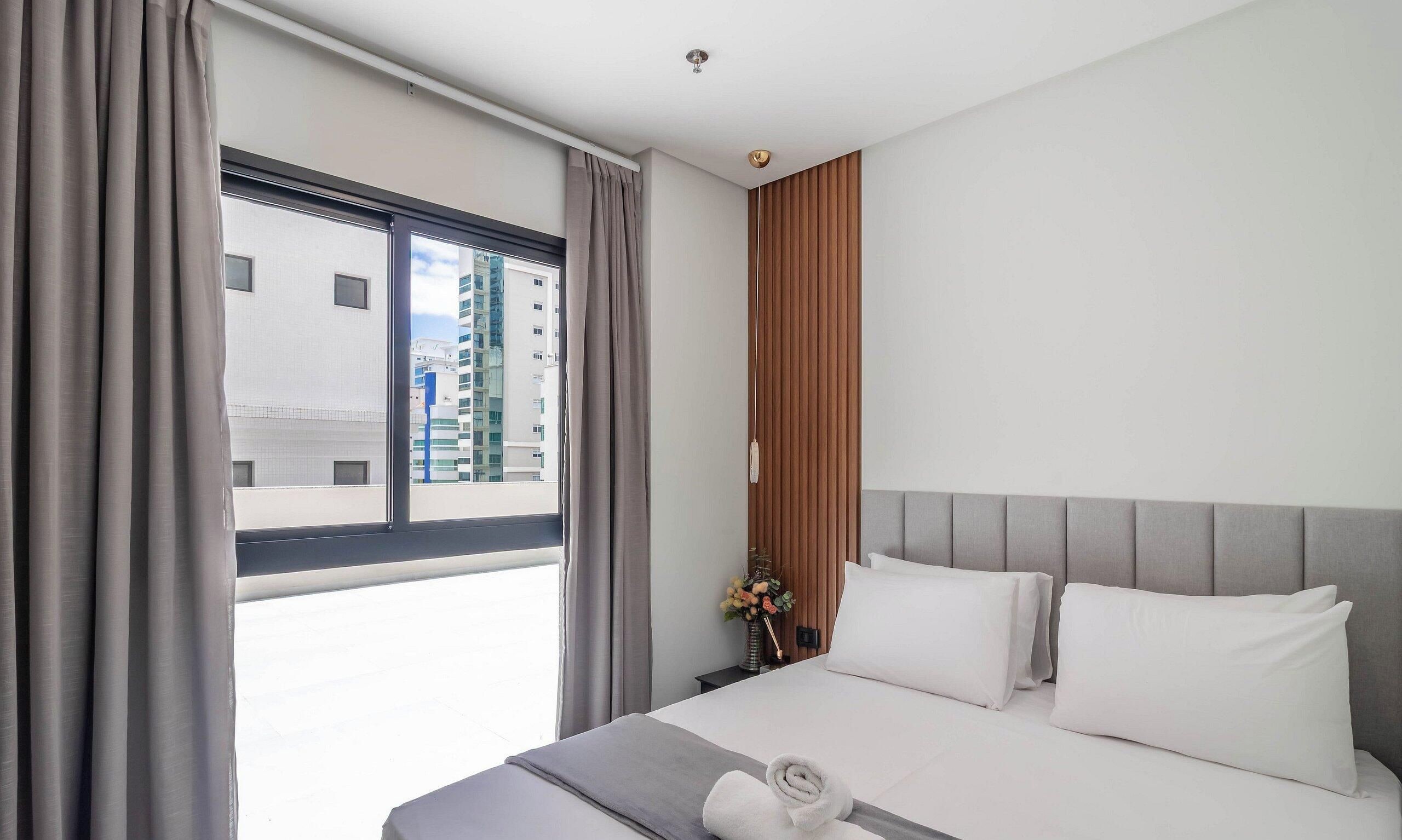 Ein Hotelzimmer mit einem großen Bett, einem Nachttisch, einem Fenster mit Vorhängen und Blick auf Gebäude außerhalb.