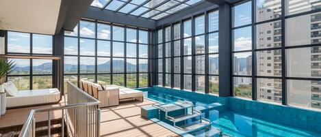 Una piscina infinita en la azotea con techo de vidrio, cubierta de madera y zona de estar con vistas a la ciudad.