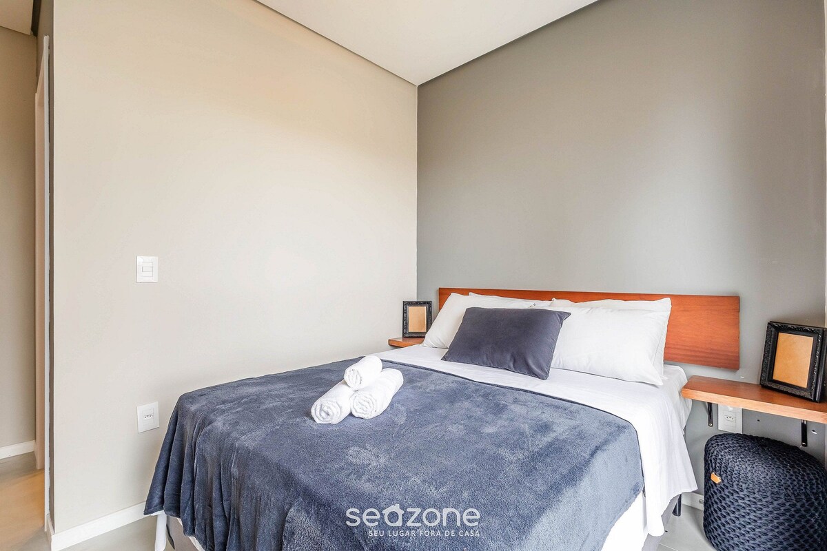 A hotel room with a bed, a bedside table, a chair, and a wall-mounted picture frame.