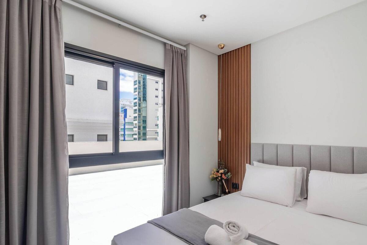 A hotel room with a bed, curtains, a nightstand, a vase with flowers, and a view of buildings through the window.