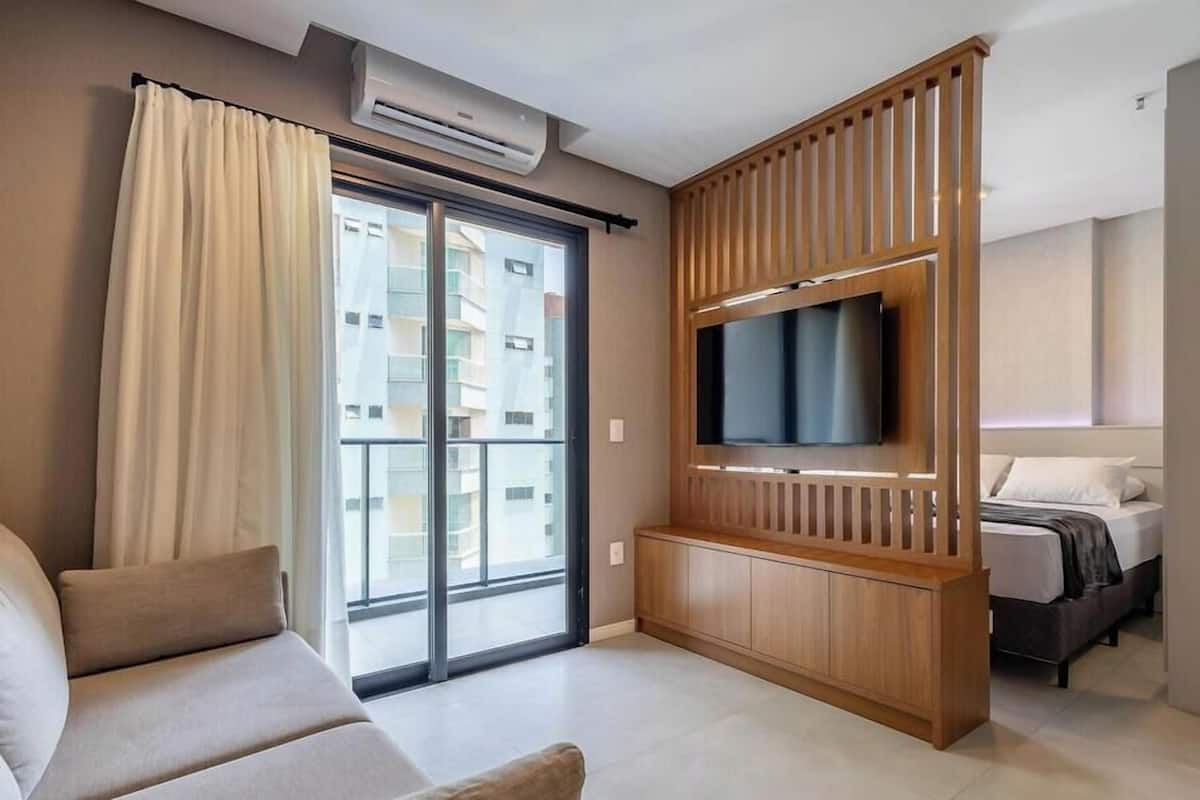 A modern hotel room with a sofa, a TV on a wooden stand, a balcony with a view of other buildings, and a bed with a white headboard.