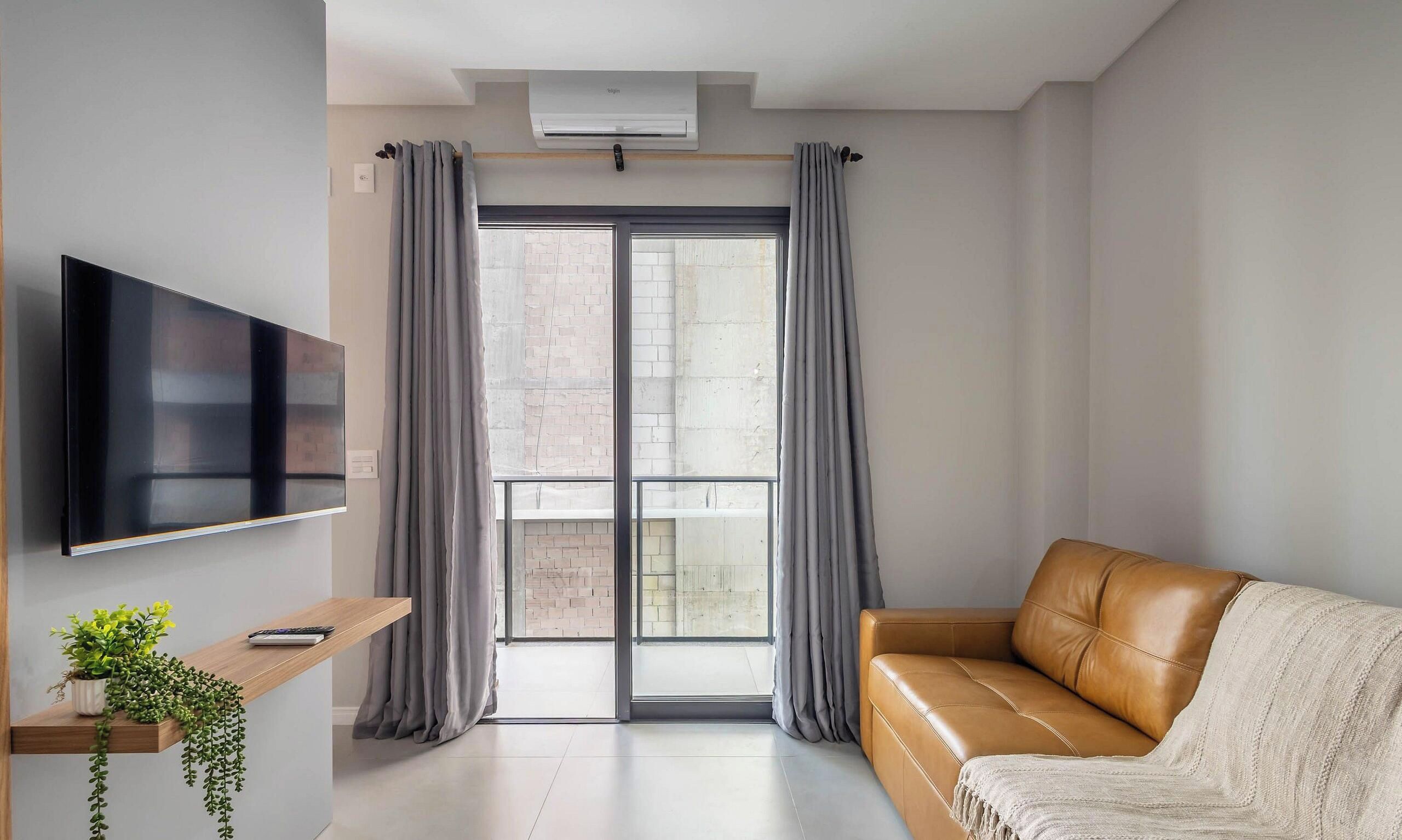 A modern living room with a flat-screen TV, a leather sofa, a small wooden table with a plant, and a sliding glass door with curtains.