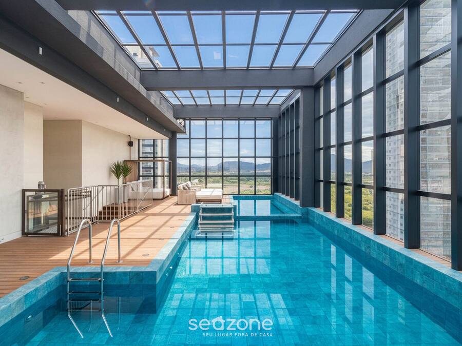 Une piscine intérieure dotée d’une terrasse en bois, d’un toit en verre et offrant une vue sur le paysage urbain.