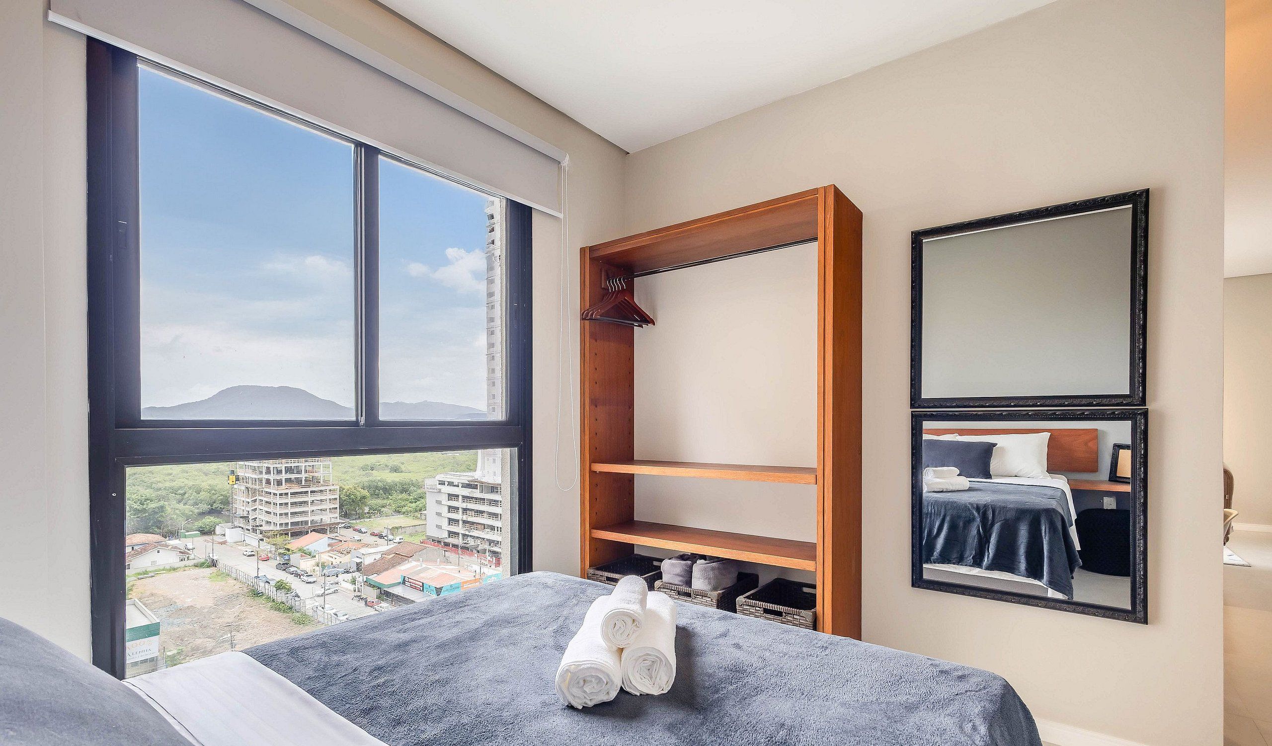 Hotellihuone, jossa on suuri ikkuna, sinisellä peitolla varustettu sänky, puinen vaatekaappi ja peili seinällä.