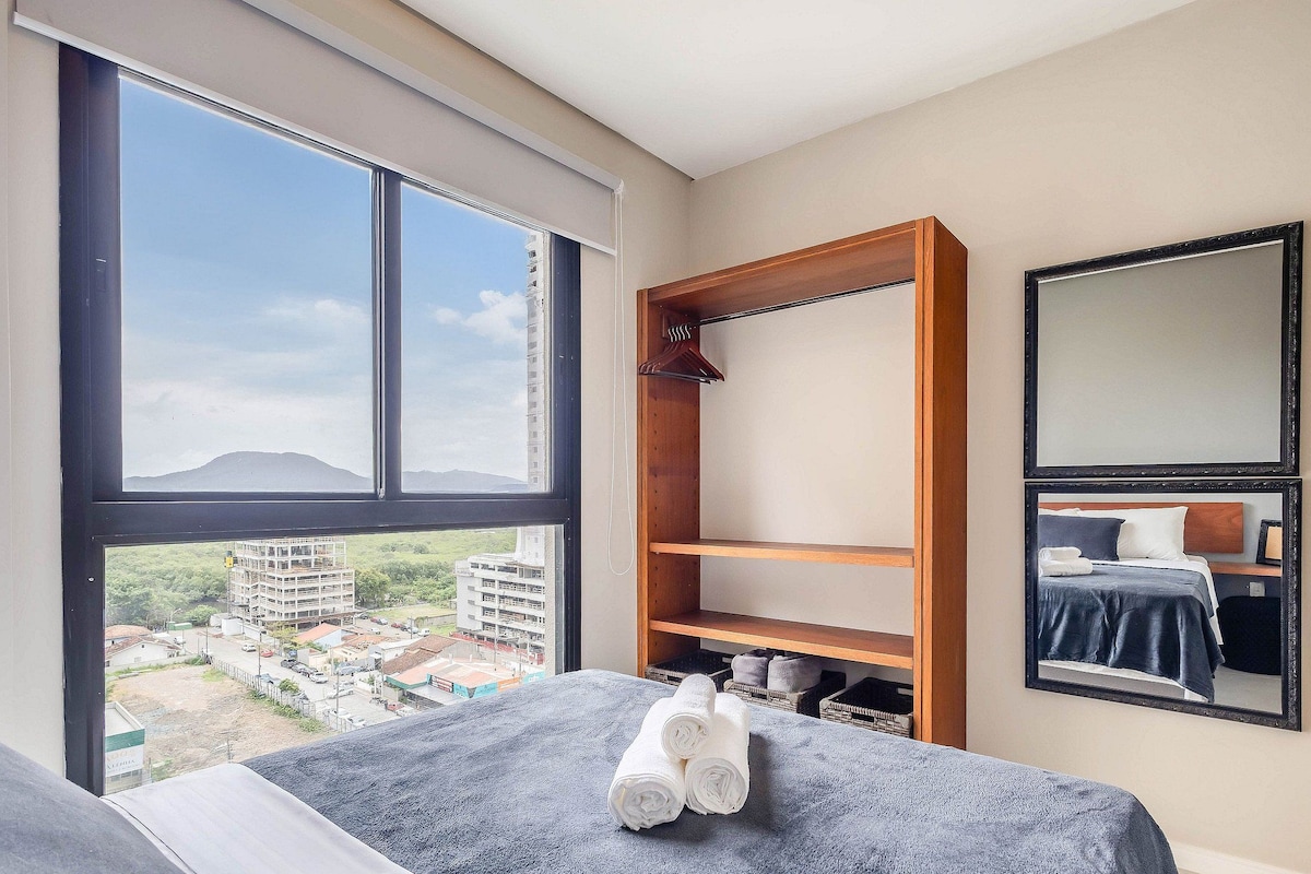 A hotel room with a large window, a bed with a blue blanket, a wooden wardrobe, and a mirror on the wall.