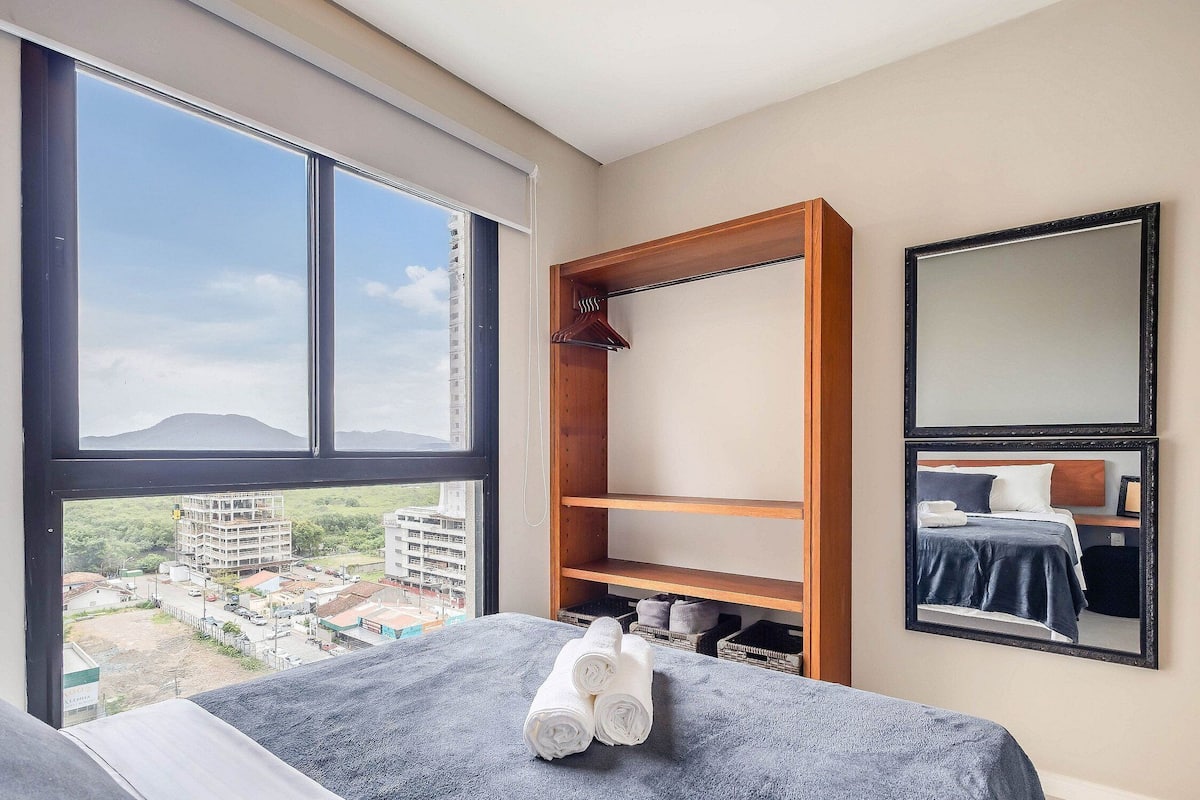 A hotel room with a large window, a bed with a blue blanket, a wooden wardrobe, and a mirror on the wall.