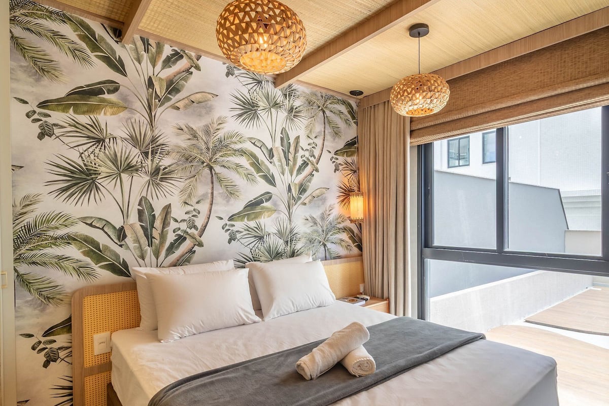 A bedroom with a large bed, tropical wallpaper, and a view of the outside.