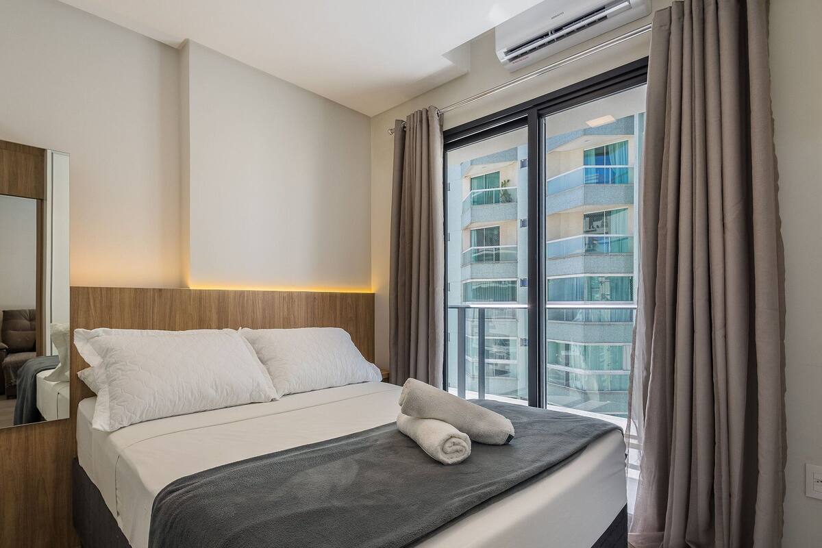 A hotel room with a bed, a mirror, an air conditioning unit, and a window with curtains.