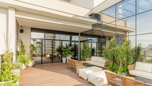 A rooftop terrace with outdoor seating, potted plants, and large windows offering city views. - Sua estadia exclusiva a poucos passos do Mar em Meia Praia - FFS (Itapema)