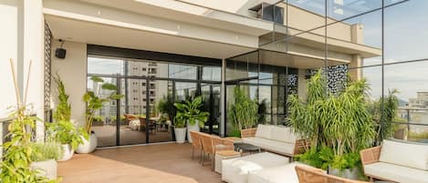 A rooftop terrace with outdoor seating, potted plants, and large windows offering city views.