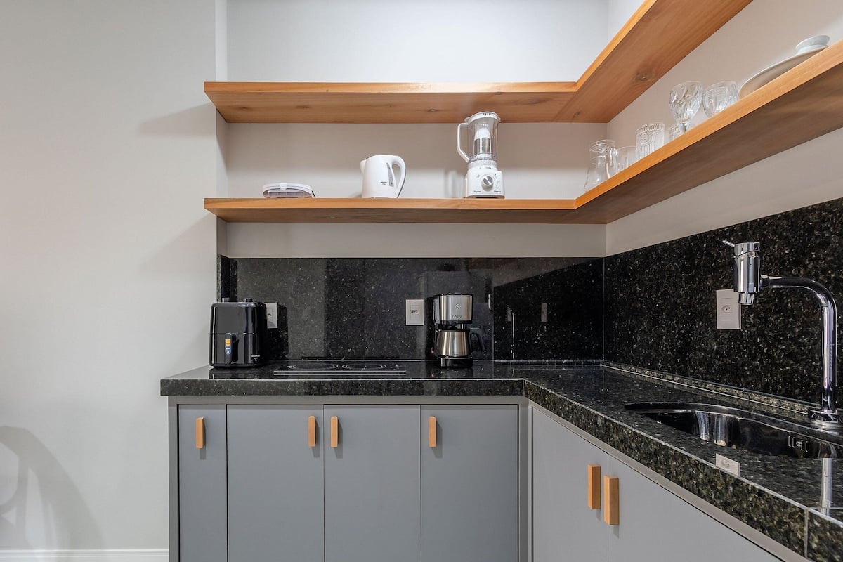 A kitchen with grey cabinets, a black countertop, a coffee maker, a blender, and a wooden shelf above.