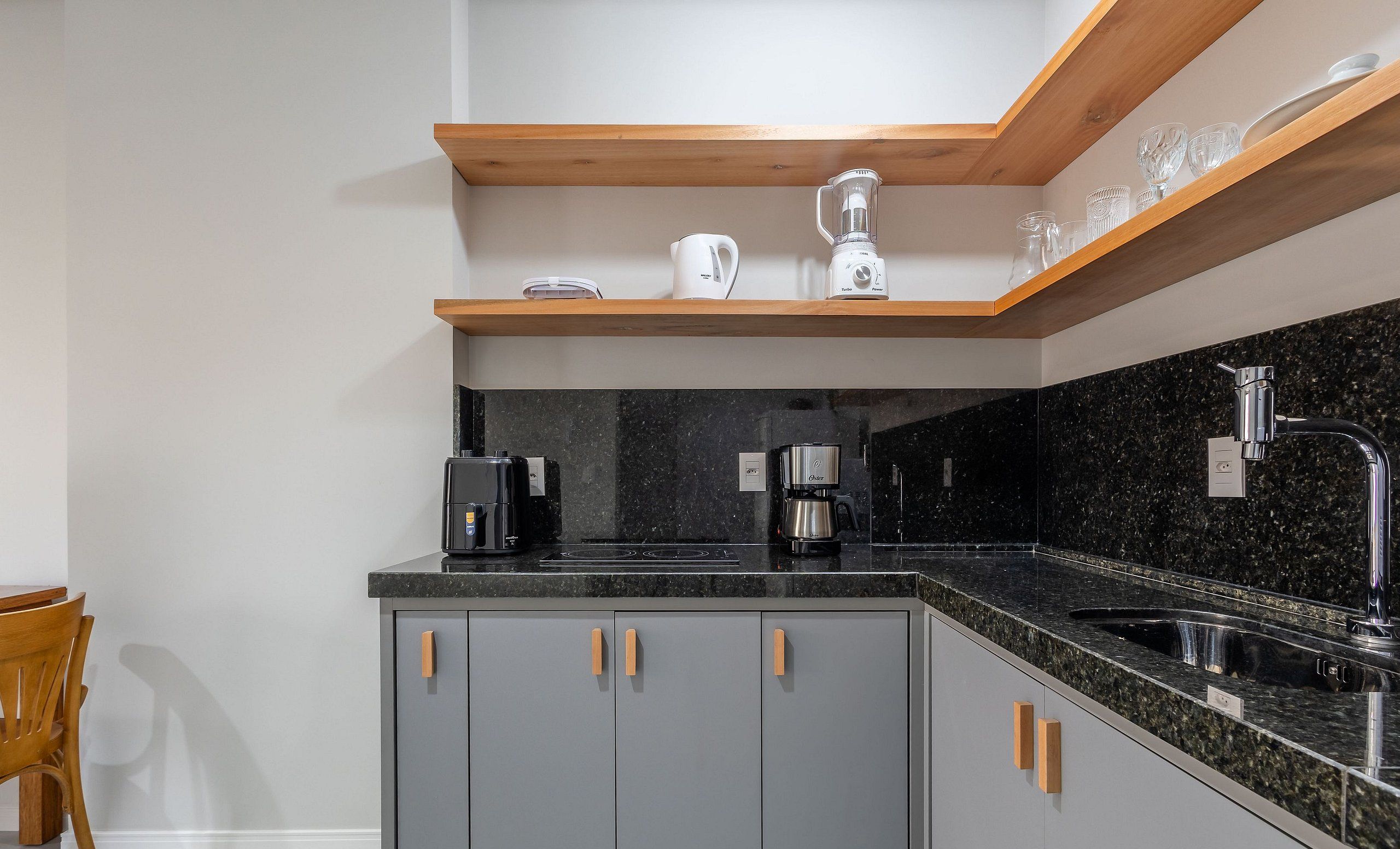 A kitchen with grey cabinets, a black countertop, a coffee maker, a blender, and a wooden shelf above.