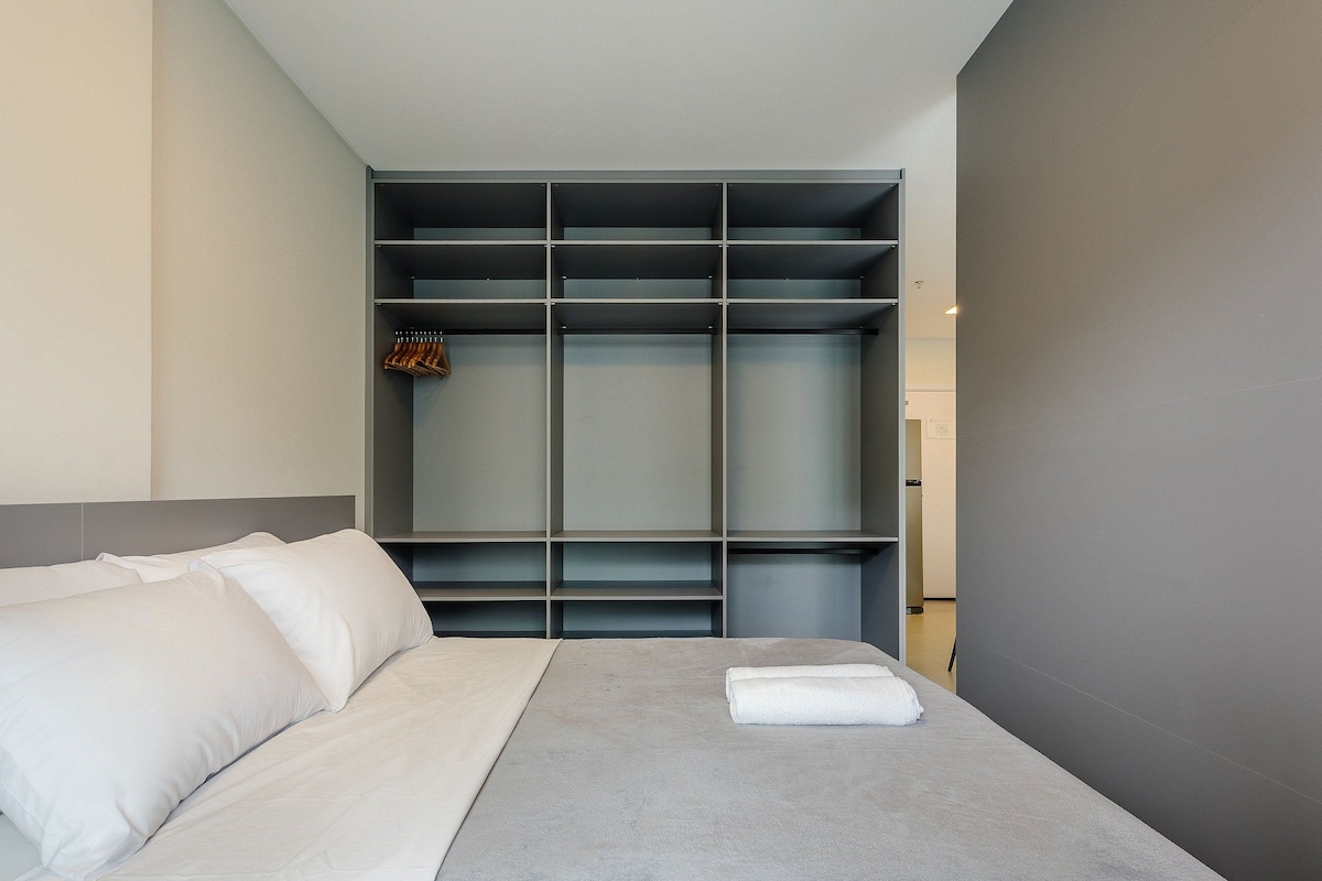 A modern hotel room with a bed, grey walls, and a built-in wardrobe with multiple shelves.