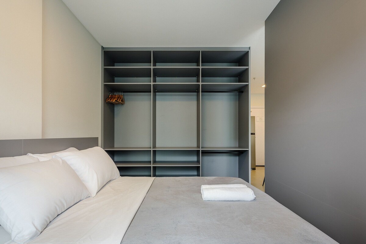 A modern hotel room with a bed, grey walls, and a built-in wardrobe with multiple shelves.