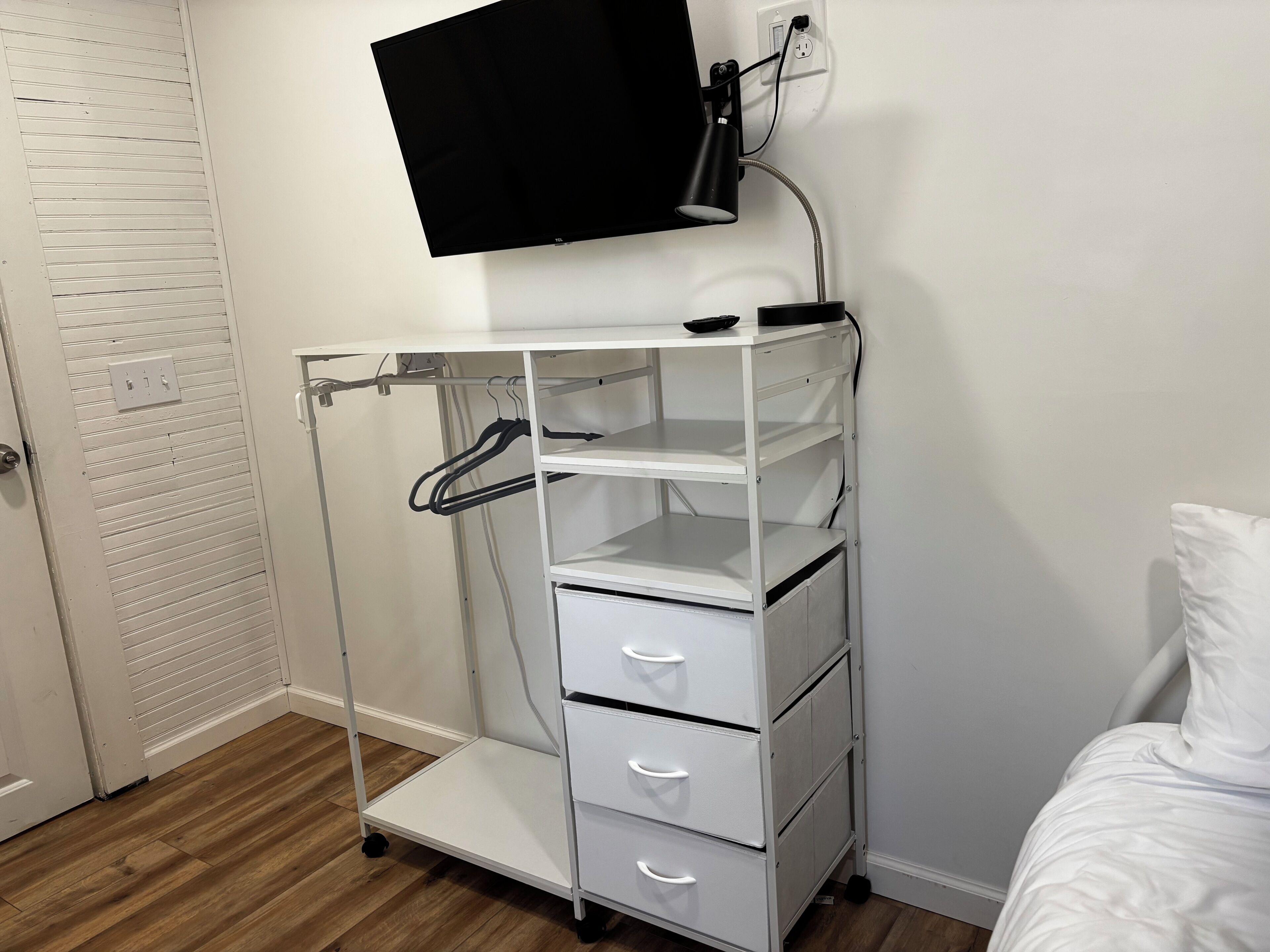 A white metal cart with drawers and shelves, a flat-screen TV mounted on the wall, and a door with a louvered design.