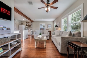 A spacious living area with a ceiling fan, a flat-screen TV, and a kitchen in the background. - Lovely cottage, downtown Bastrop Historic District (Bastrop)