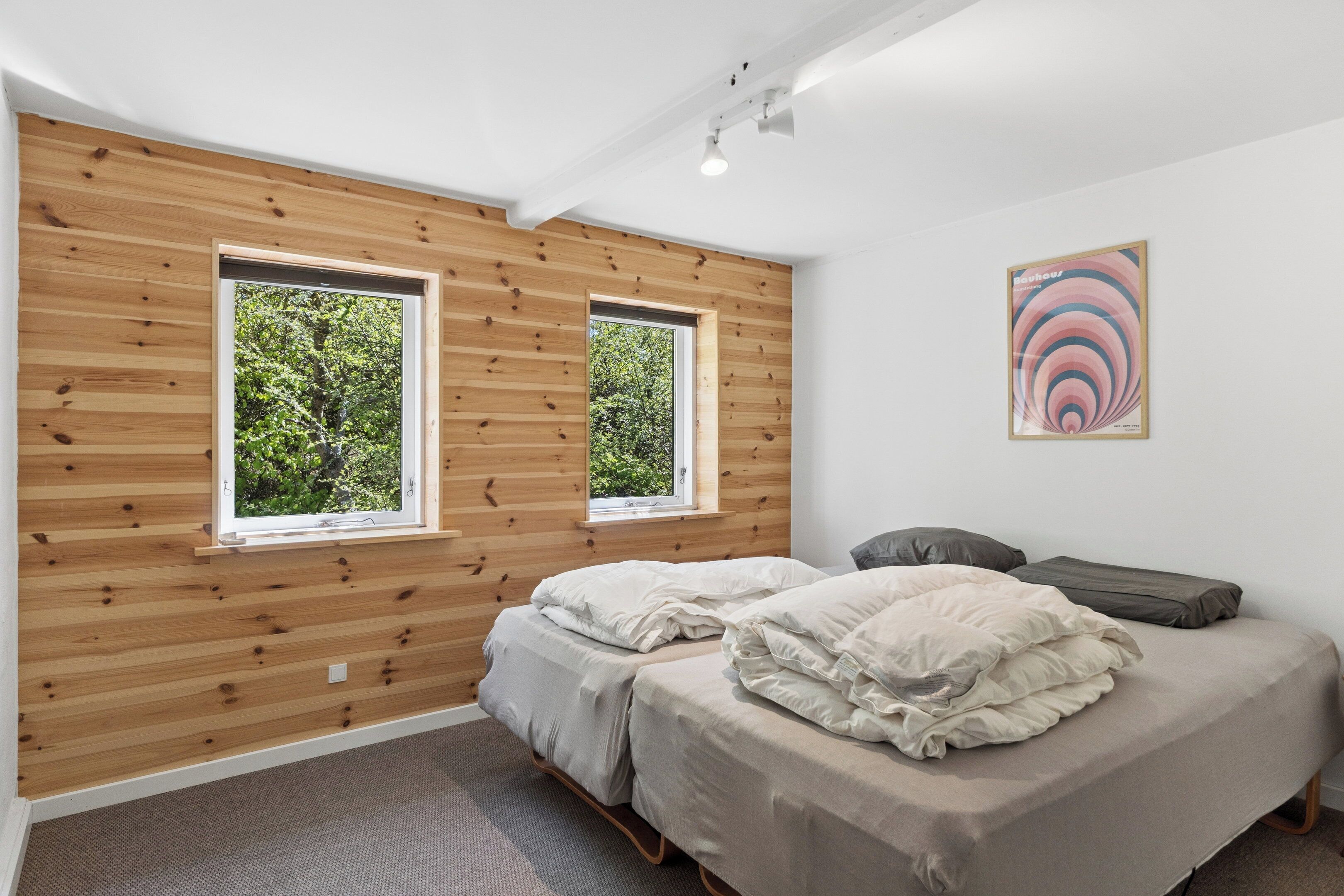 A bedroom with two beds, wooden panel walls, and two windows with a view of greenery.