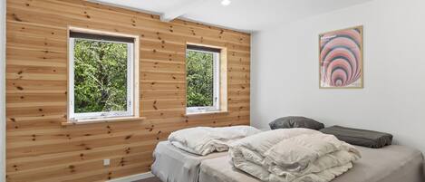 A bedroom with two beds, wooden panel walls, and two windows with a view of greenery.