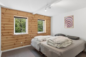 A bedroom with two beds, wooden panel walls, and two windows with a view of greenery.