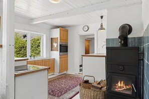 A modern kitchen with white cabinetry, a built-in oven, a stove with a visible fire, a clock on the wall, and a view of greenery outside.