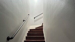 Una escalera de madera con barandillas metálicas, que lleva a un rellano con una fuente de luz en la parte superior.