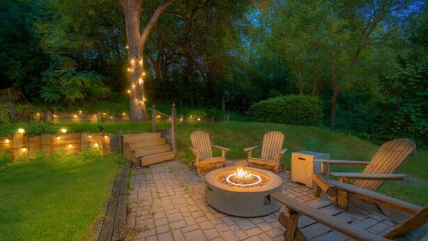 Un hogar de fuego rodeado de sillas Adirondack de madera en un patio empedrado, con luces colgantes y una cerca de madera en un patio trasero arbolado.