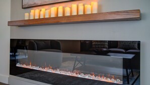 Una chimenea moderna con frente de vidrio, un estante de madera encima con velas encendidas, y un cuadro abstracto enmarcado en la pared.
