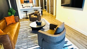 Un salón moderno con un sofá de cuero marrón, un sillón gris y una mesa de centro. Un televisor grande está montado en la pared, y se ve una cocina al fondo.