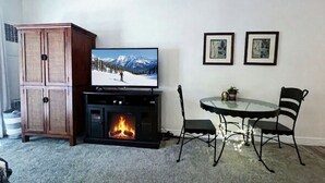 Un salon avec une cheminée, un téléviseur, une table à manger avec des chaises et deux tableaux encadrés au mur.