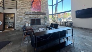Un espace intérieur vaste et lumineux, doté d’une cheminée en pierre, d’une grande fenêtre et d’un coin salon avec des tables et des chaises.