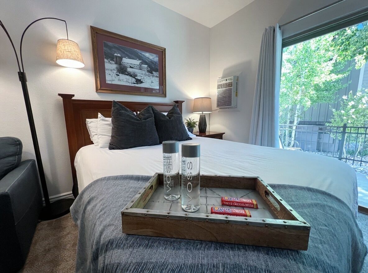 Een hotelkamer met een bed, nachtlampjes, een ingelijst schilderij aan de muur, en een raam met uitzicht op bomen.