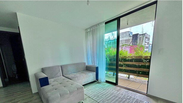 Un salon moderne avec un canapé d’angle gris, un petit pouf et une porte vitrée donnant sur un balcon extérieur.