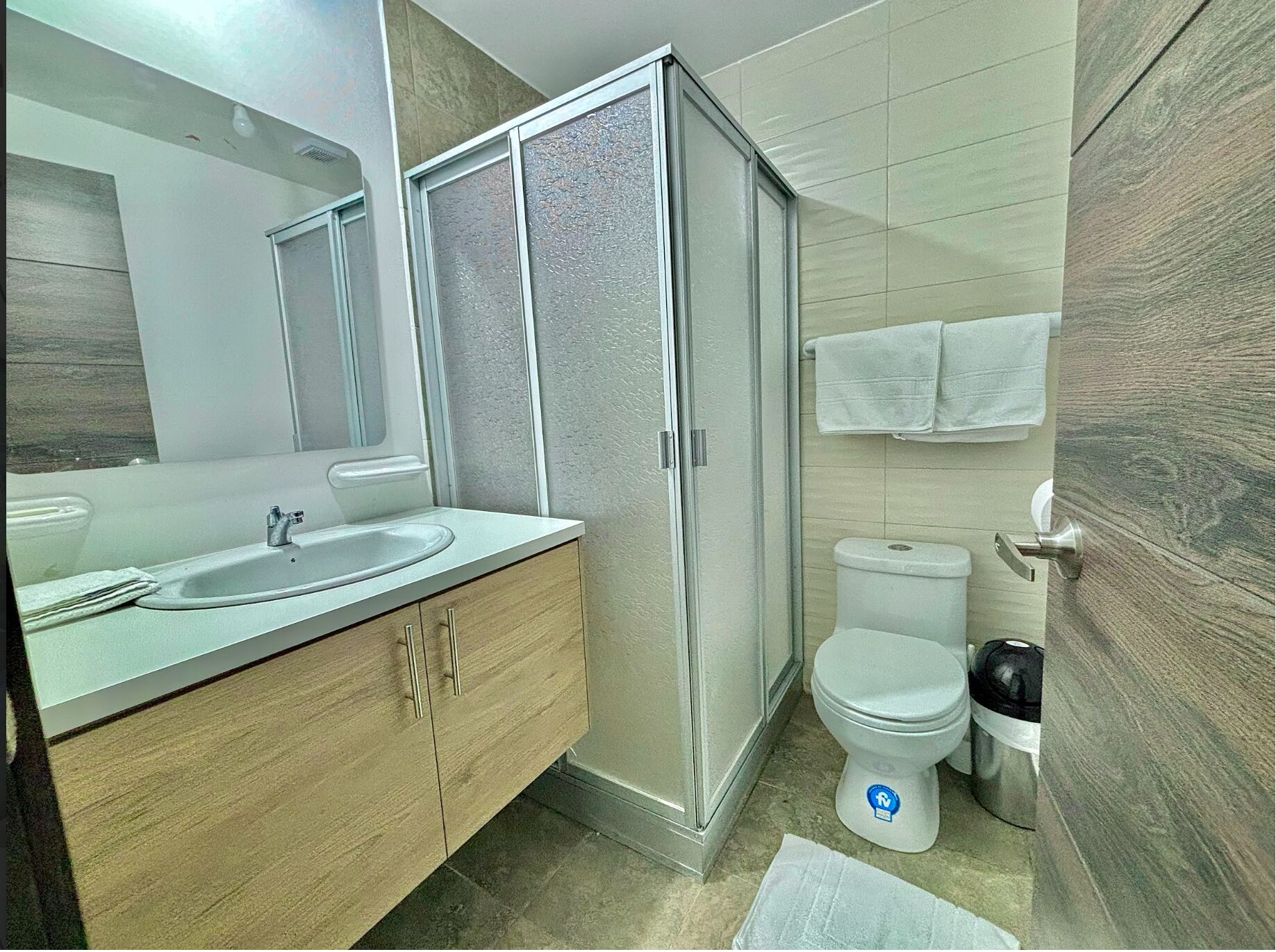 Un baño moderno con cabina de ducha de vidrio, lavamanos con espejo, inodoro y toallero.