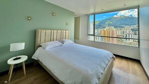 A bedroom with a bed, a bedside table with a lamp, and a large window with a cityscape view. - Luxury 2BR + Gym & City Views| Near Spain Embassy (QUITO)