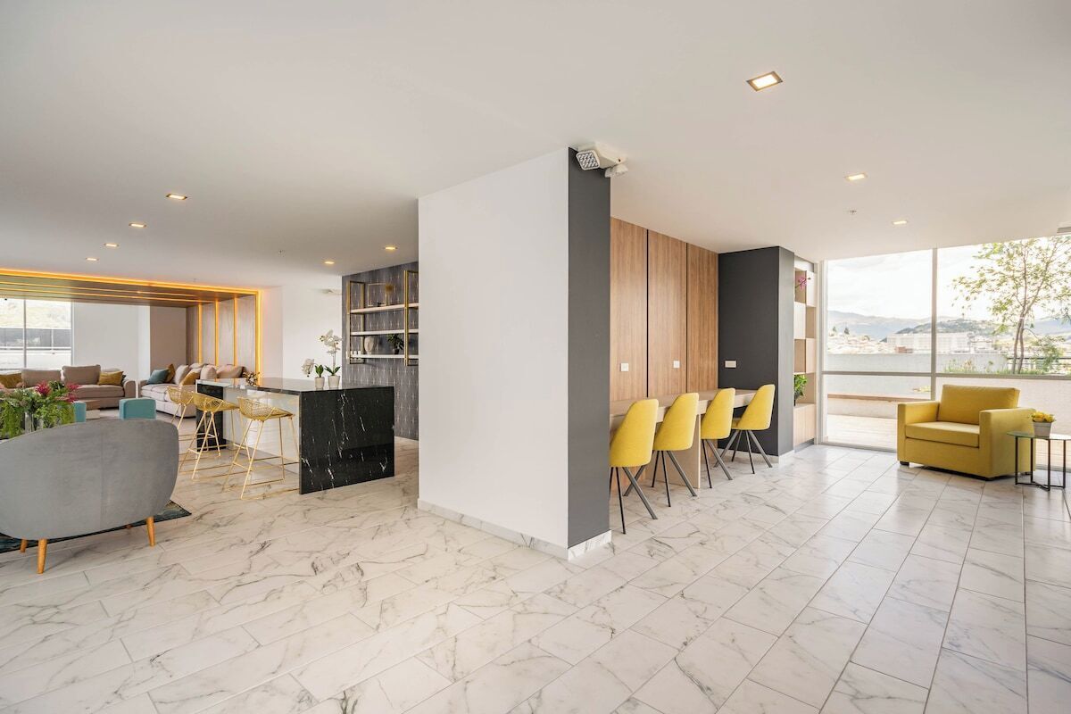A modern living room with a sofa, a dining area with yellow chairs, and a kitchen with a black countertop.