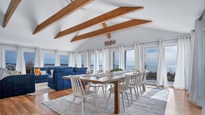 Un vaste espace de vie doté d’un plafond en bois, d’une table à manger avec des chaises, d’un canapé bleu et de grandes fenêtres offrant une vue sur la mer.