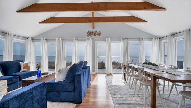 Un vaste espace de vie doté d’un haut plafond, de grandes fenêtres, d’un canapé bleu, d’une table à manger en bois et d’une vue sur la mer.