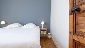 Un dormitorio con una cama, dos lámparas de noche, una mesita de noche de madera con una lámpara, un estante pequeño con libros y una puerta de madera.