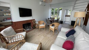 Una sala de estar con chimenea, sillas de mimbre, un sofá con cojines, un televisor y un comedor con sillas.