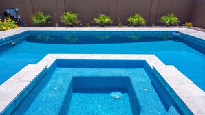 A rectangular swimming pool with a shallow end and a deeper section, surrounded by a tiled deck and small plants.