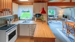 Een moderne keuken met houten kastjes, een ingebouwde oven en een groot kookeiland met spoelbak. Er is een eethoek met een tafel en stoelen, en een rode hanglamp erboven.